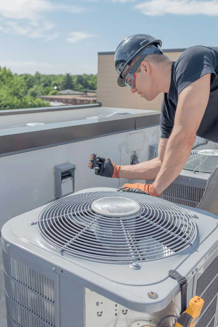A technician in safety gear performs AC repair on a rooftop air conditioning unit, adjusting controls on a nearby panel under a clear sky.