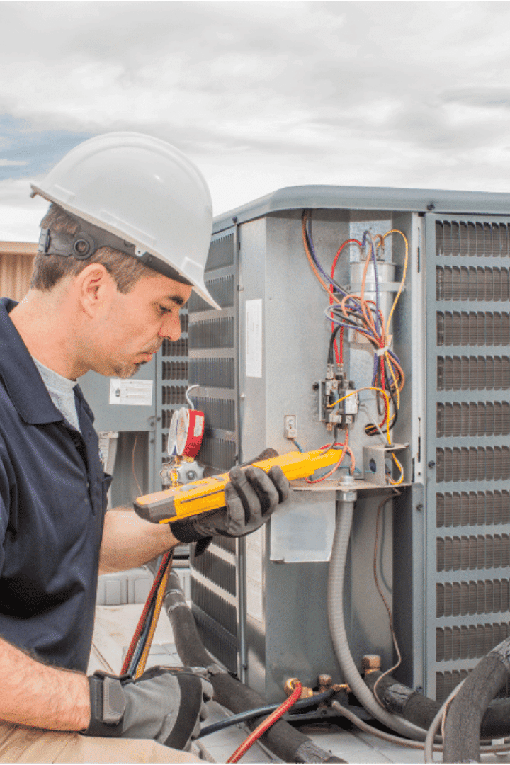 A technician wearing a hard hat and gloves uses a multimeter to inspect the wiring of an outdoor air conditioning unit during an ac repair.