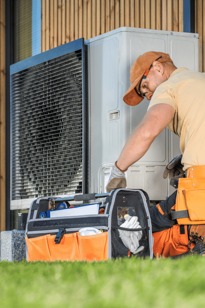A technician wearing an orange cap and tool belt performs ac repair on an outdoor air conditioning unit, with an open tool bag on the grass beside him.