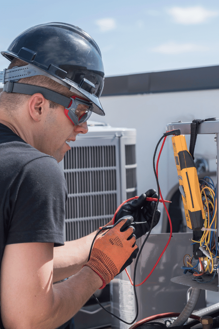 A technician wearing safety gear uses a multimeter to test electrical connections on HVAC equipment outdoors during an ac repair.