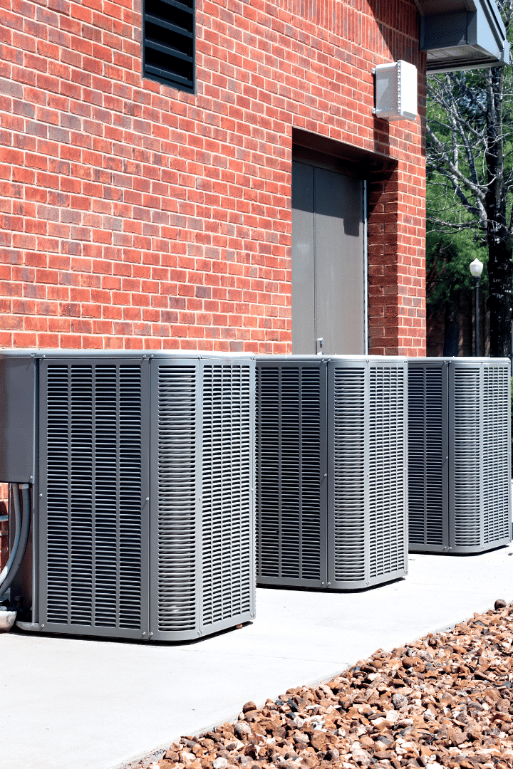 Three large outdoor air conditioning units, ready for any ac repair needs, are installed on a concrete pad next to a red brick building.