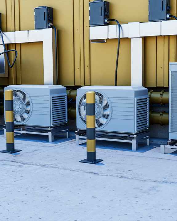 Three outdoor air conditioning units are installed on concrete near a building wall, with two yellow and black safety bollards positioned in front—an easy-access setup for future ac repair needs.